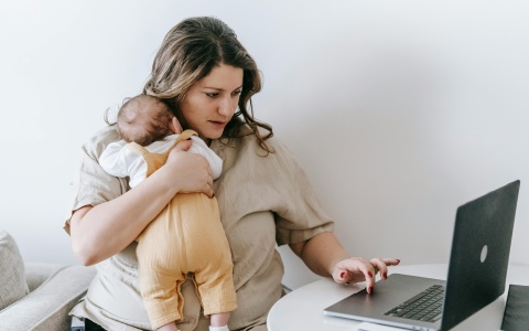Eine Frau hält ein Baby auf dem Arm, mit der anderen Hand bedient sie ihren Laptop.
