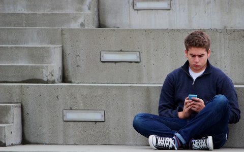 Ein Junge im Teenageralter sitzt im Schneidersitz auf einer grauen Treppe und sieht bedrückt auf sein Smartphone.