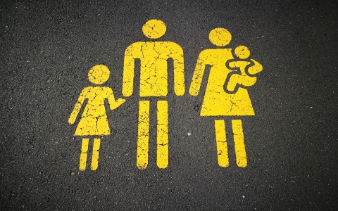 Auf einem Asphaltboden ist in gelber Farbe das Piktogram einer Familie aufgebracht. Der Vater hält eine Tochter an der Hand und die Frau trägt ein Kleinkind.