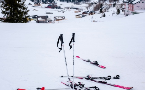 Drei Paar Skier und Skistöcke liegen im Schnee. Im Hintergrund sind Hütten und schneebedeckte Berge zu sehen.