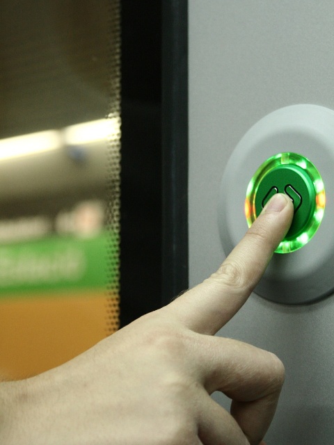 Das Bild zeigt einen Finger, der den Button einer automatischen Tür in einem Öffentlichen Verkehrsmittel drückt.
