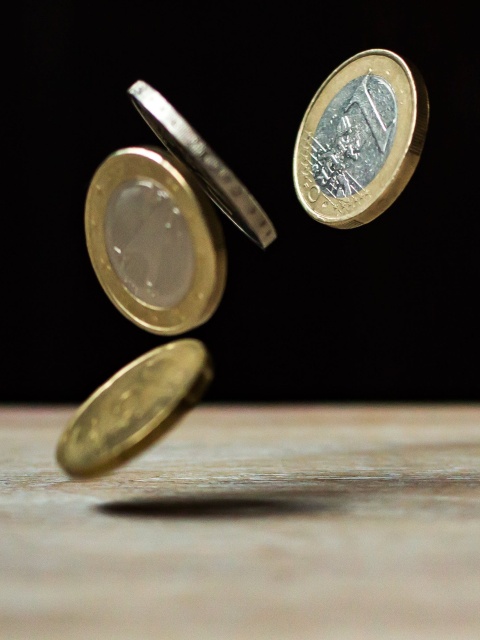 Mehrere Euromünzen fallen auf einen Tisch. Der Hintergrund ist schwarz.