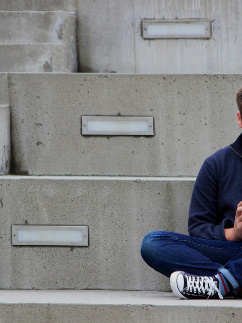 Ein Junge im Teenageralter sitzt im Schneidersitz auf einer grauen Treppe und sieht bedrückt auf sein Smartphone.